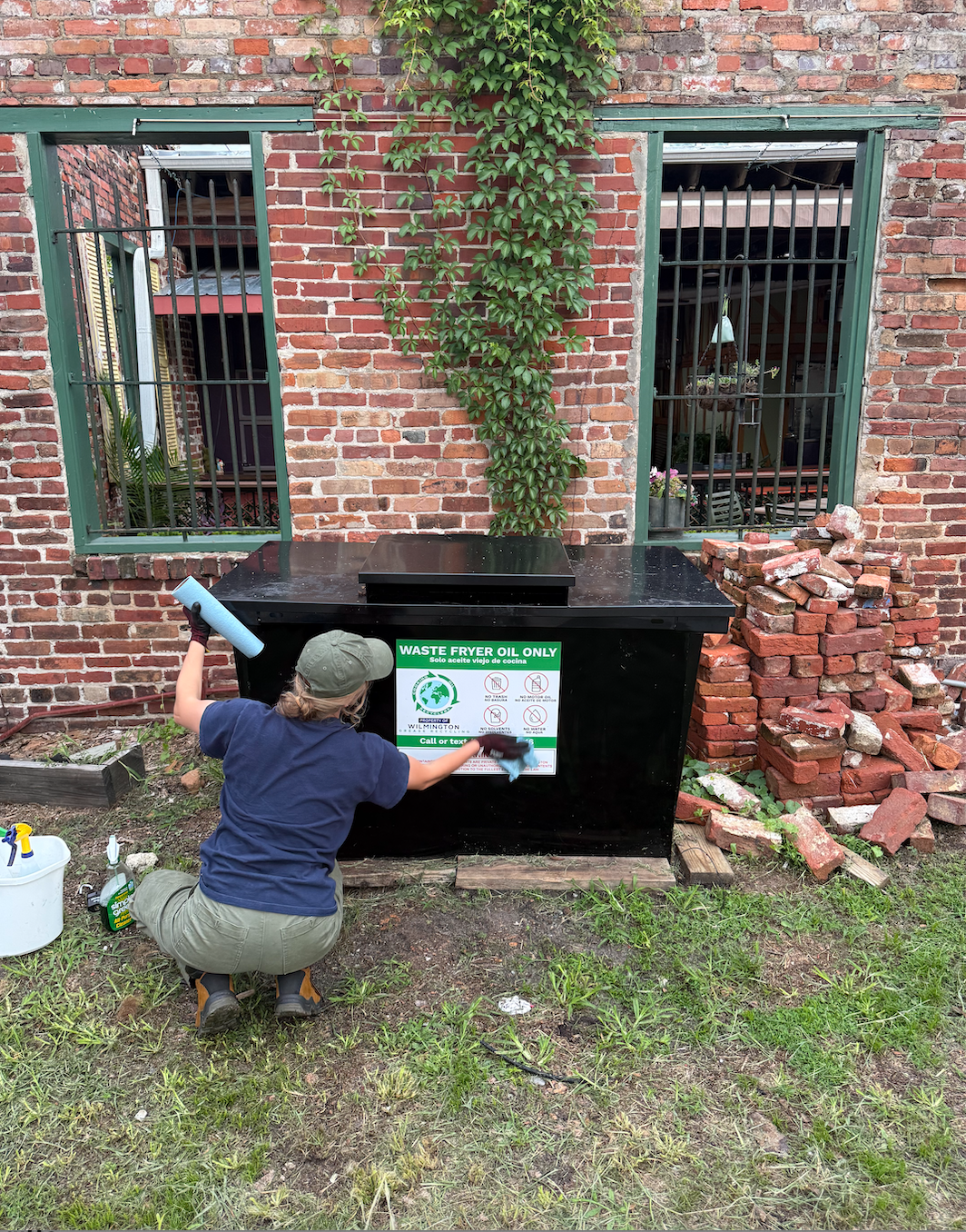 Worker installing a waste fryer oil recycling container outside a brick commercial building.